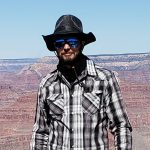 Scott Smith in a full brimmed hat, sunglasses and a flannel standing on the rim of the Grand Canyon