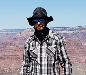 Scott Smith in a full brimmed hat, sunglasses and a flannel standing on the rim of the Grand Canyon