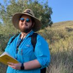 Liam Schramm standing in a grassy field holding a yellow notebook wearing a full brimmed hat, sun glasses and a blue shirt