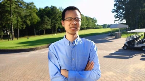 Zhifeng Hu wearing a blue button up shirt and glasses with his arms crossed standing in front of a wooded area