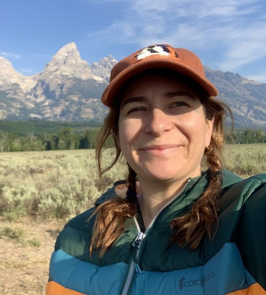 Christine Sestak wearing a billed hat and puffy vest with the Tetons in the background