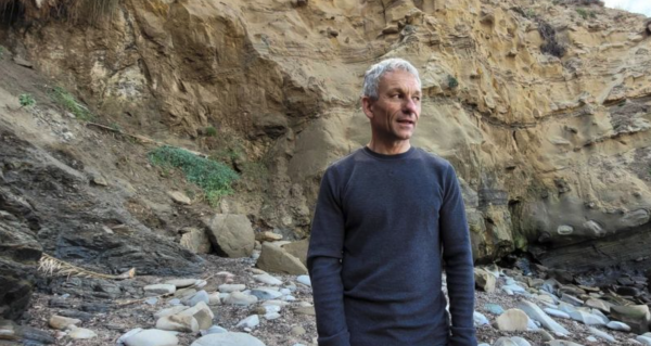 A man wearing a navy long sleeve shirt stands on a rocky beach looking at the ocean