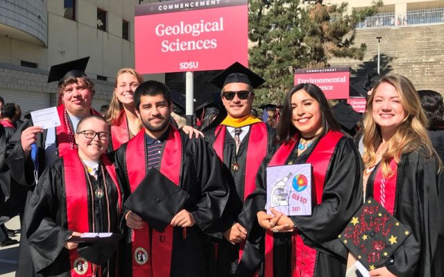 Undergraduates and Graduates wearing graduation regalia gathered together in front of the Geological Sciences sign