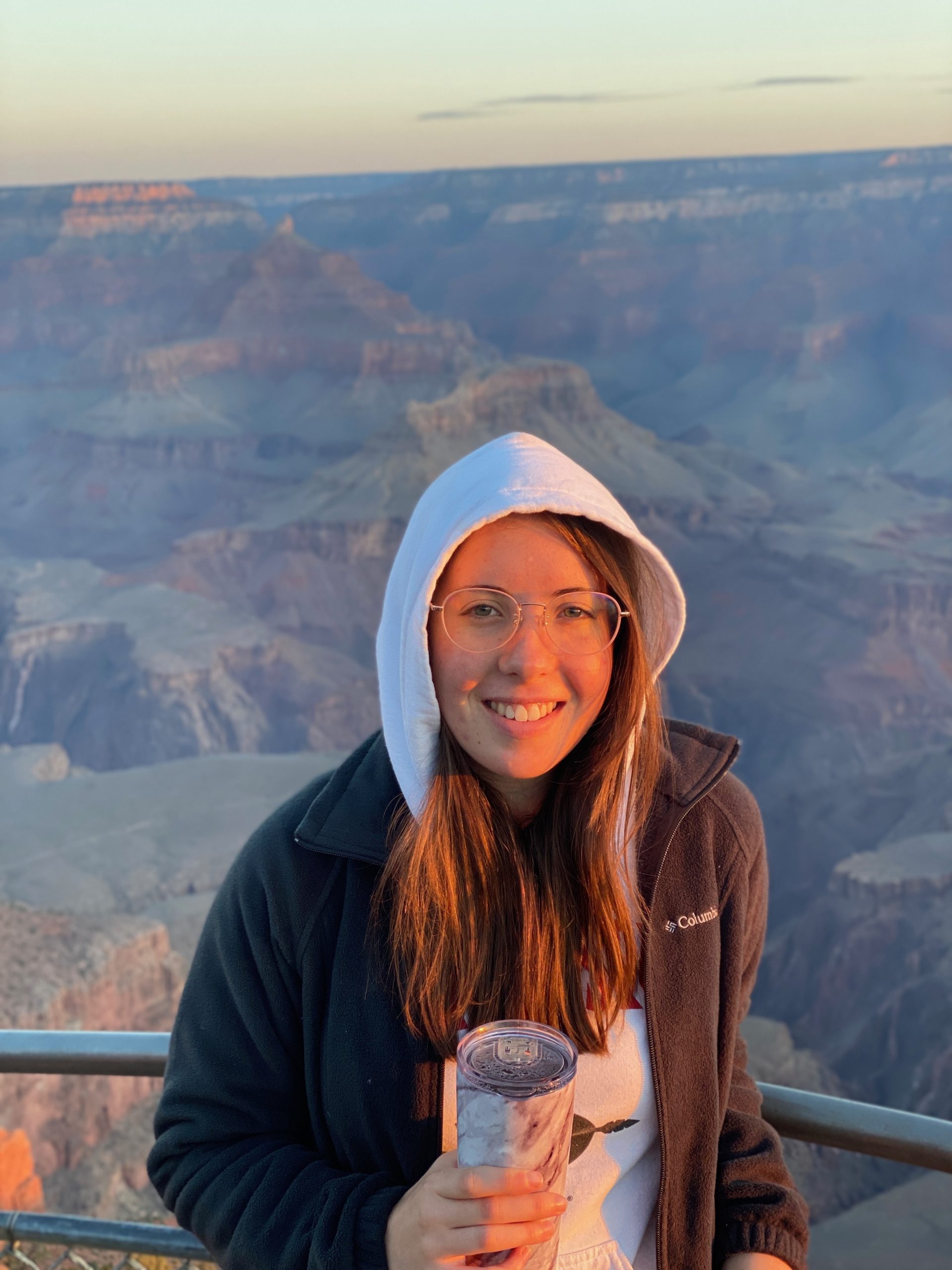 Caitlin O'Keefe wearing a white hood and glasses holding a cup in front of the Grand Canyon