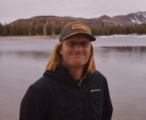 Logan Platt wearing a baseball hat and black jacket, standing in front of a lake