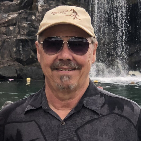 Dr. Tom Rockwell wearing a black button up collard shirt, tan baseball hat and black aviator style sunglasses