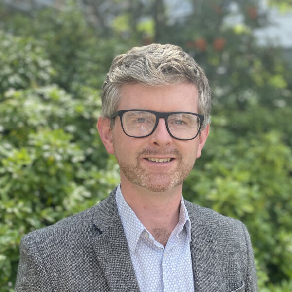 Dr. Tom Gernon wearing black rimmed glasses and a grey and white tweed suit jacket over a grey button up collard shirt