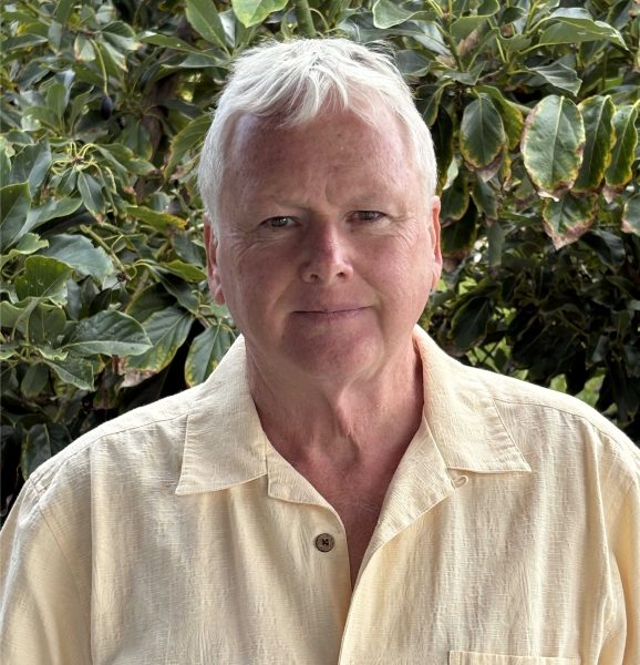 Danny Sims wearing pale yellow collared shirt in front of green leafs